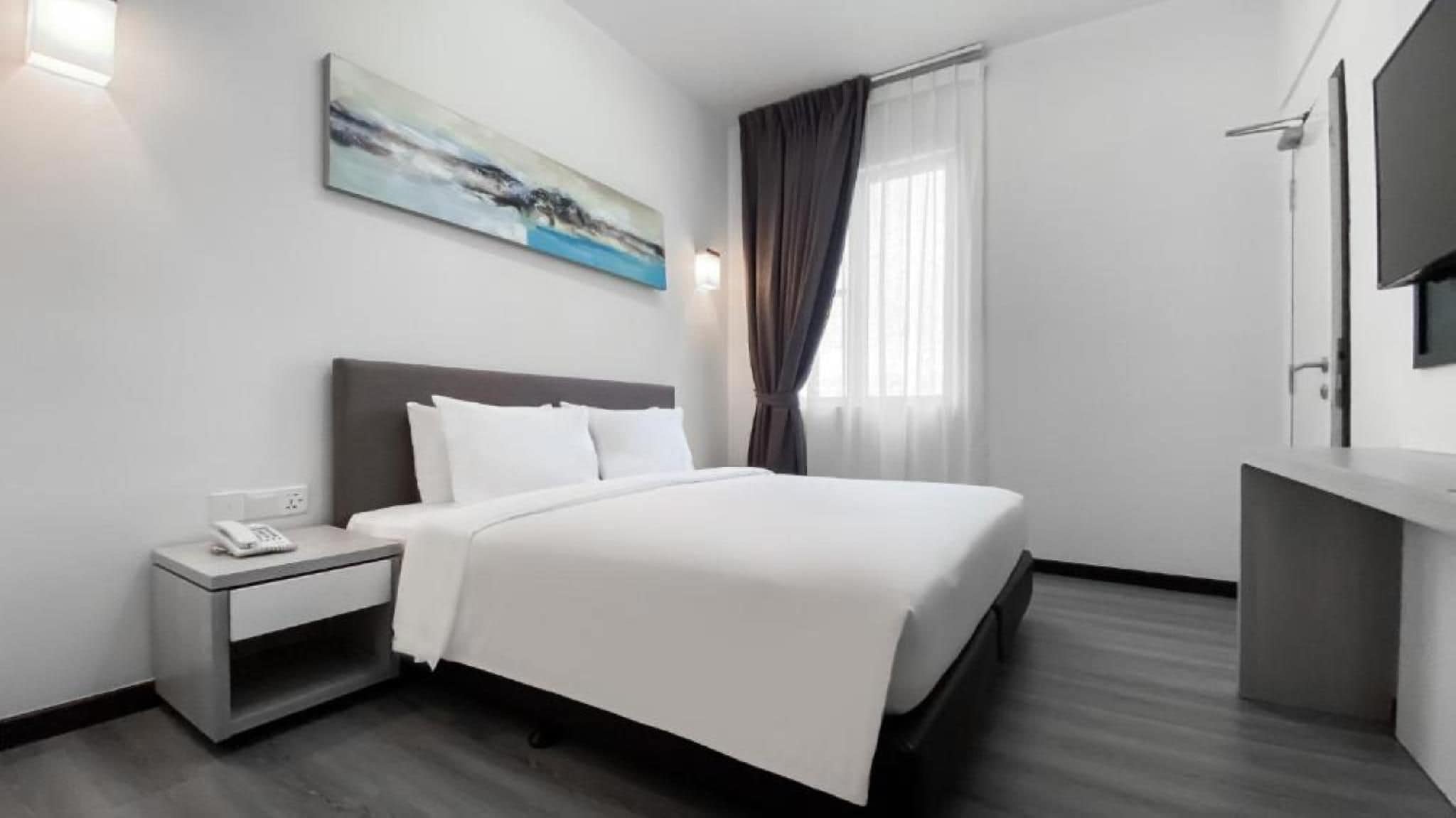 A hotel room with a bed, bedside table, a painting, a window with curtains, and a TV on the wall.