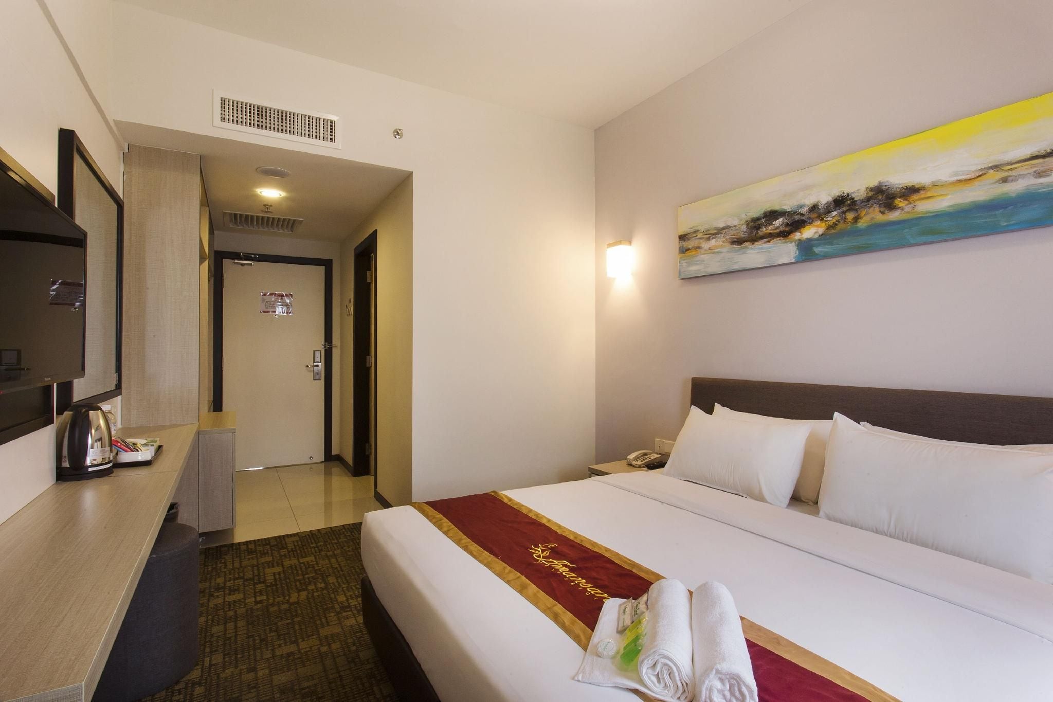 A hotel room with a bed, a desk with a kettle, a painting on the wall, and a door leading to another room.