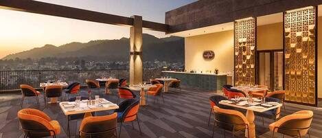 An outdoor dining area with tables and chairs, a bar, and a view of a cityscape.