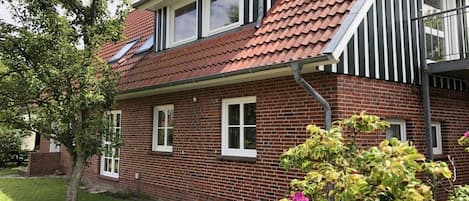 Un bâtiment en briques de deux étages, avec un toit recouvert de tuiles rouges, des fenêtres à cadre blanc et un petit jardin agrémenté d’arbres et de plantes à fleurs.