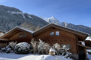 A wooden chalet with a sloped roof, surrounded by snow-covered trees and mountains in the background.