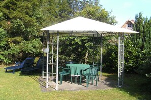 Ein überdachter Außenbereich mit Tisch und Stühlen, umgeben von viel Grün.
