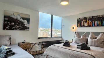 A hotel room with two beds, a large window, a city view, and a surfboard collection on the wall.