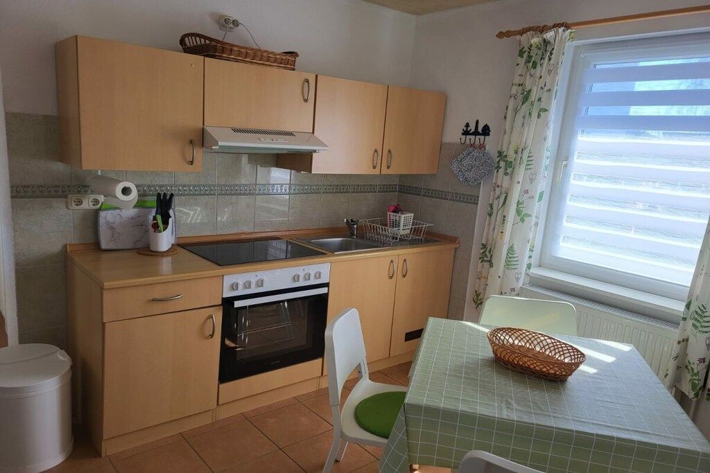 Une cuisine compacte équipée d’une cuisinière, d’un four et d’un évier, une table à manger avec un panier, et une fenêtre avec des rideaux.