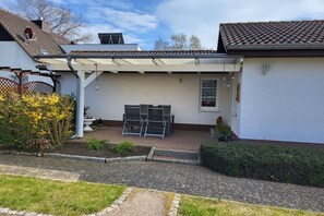 A covered patio area with a pergola, a seating arrangement, and a small garden with yellow flowers. - Holiday home with 1 bedroom (Ostseebad Rerik)