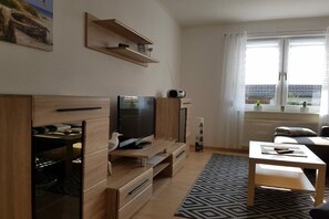A modern living room with a TV, wooden furniture, a patterned rug, and a window with curtains. - Holiday home with 1 bedroom (Ostseebad Rerik)