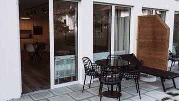 Une terrasse extérieure avec une table ronde et des chaises, un banc en bois et une porte-fenêtre coulissante donnant sur un espace intérieur comprenant une cuisine et un coin repas.