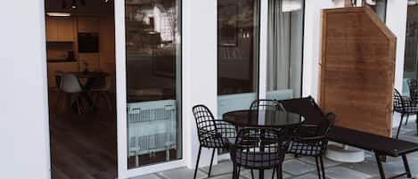An outdoor patio with a round table and chairs, a wooden bench, and a sliding glass door leading to an interior space with a kitchen and dining area.