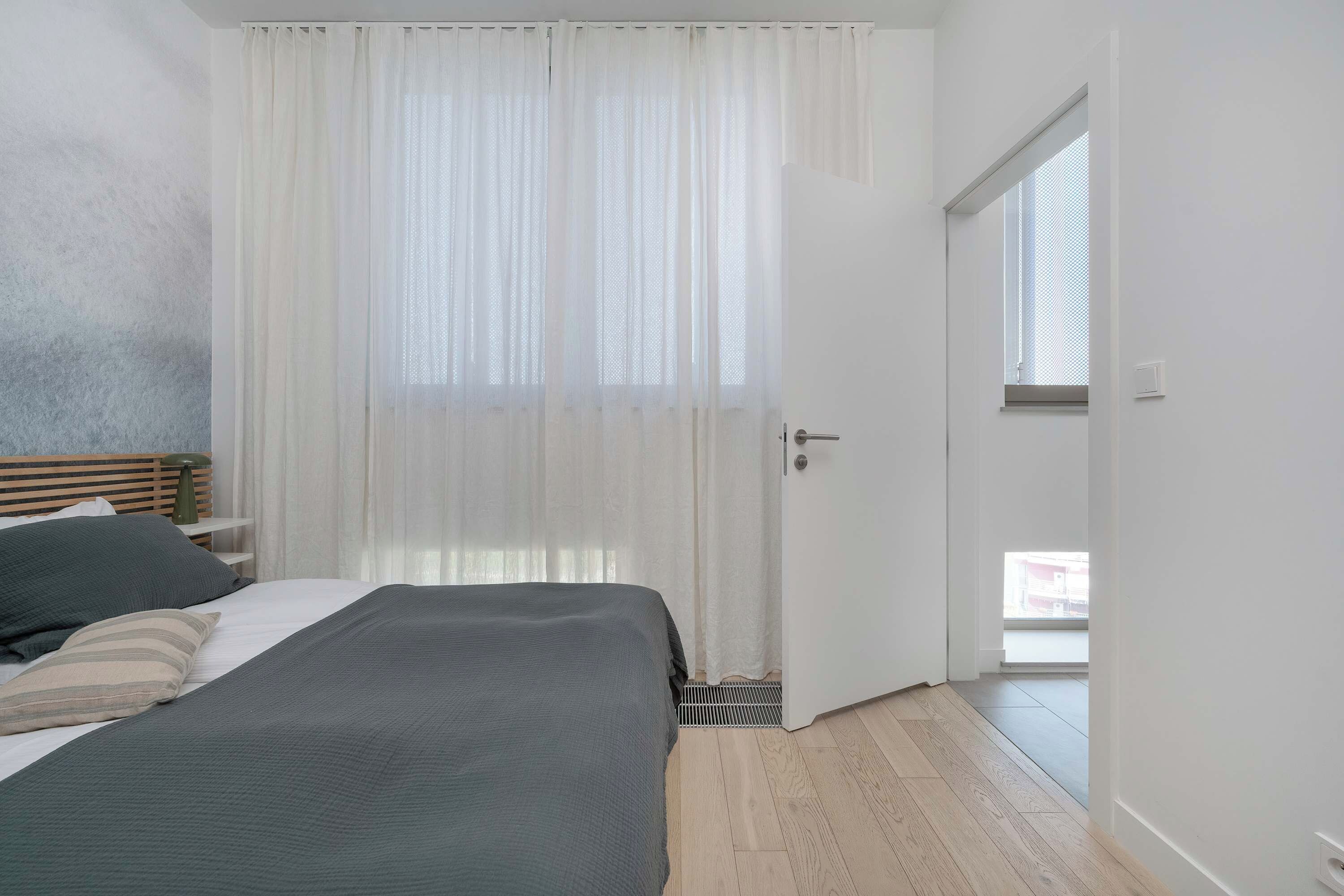Ein Schlafzimmer mit einem Bett, einem Nachttisch, einer Lampe, einem Fenster mit durchsichtigen Vorhängen und einer Tür zu einem anderen Raum.