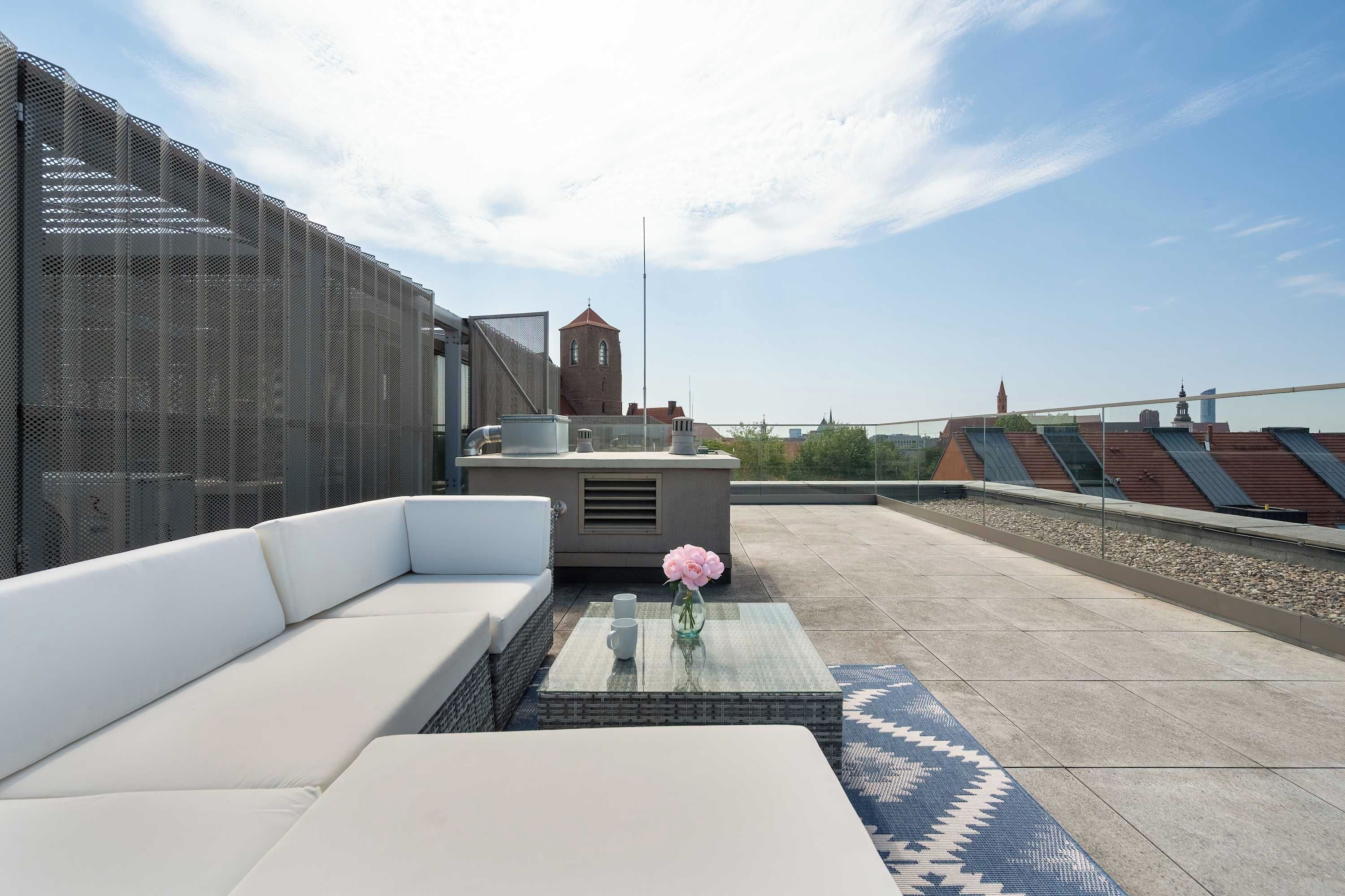 Una terraza en la azotea con un sofá blanco, una pequeña mesa con un florero y vistas a un edificio histórico al fondo.