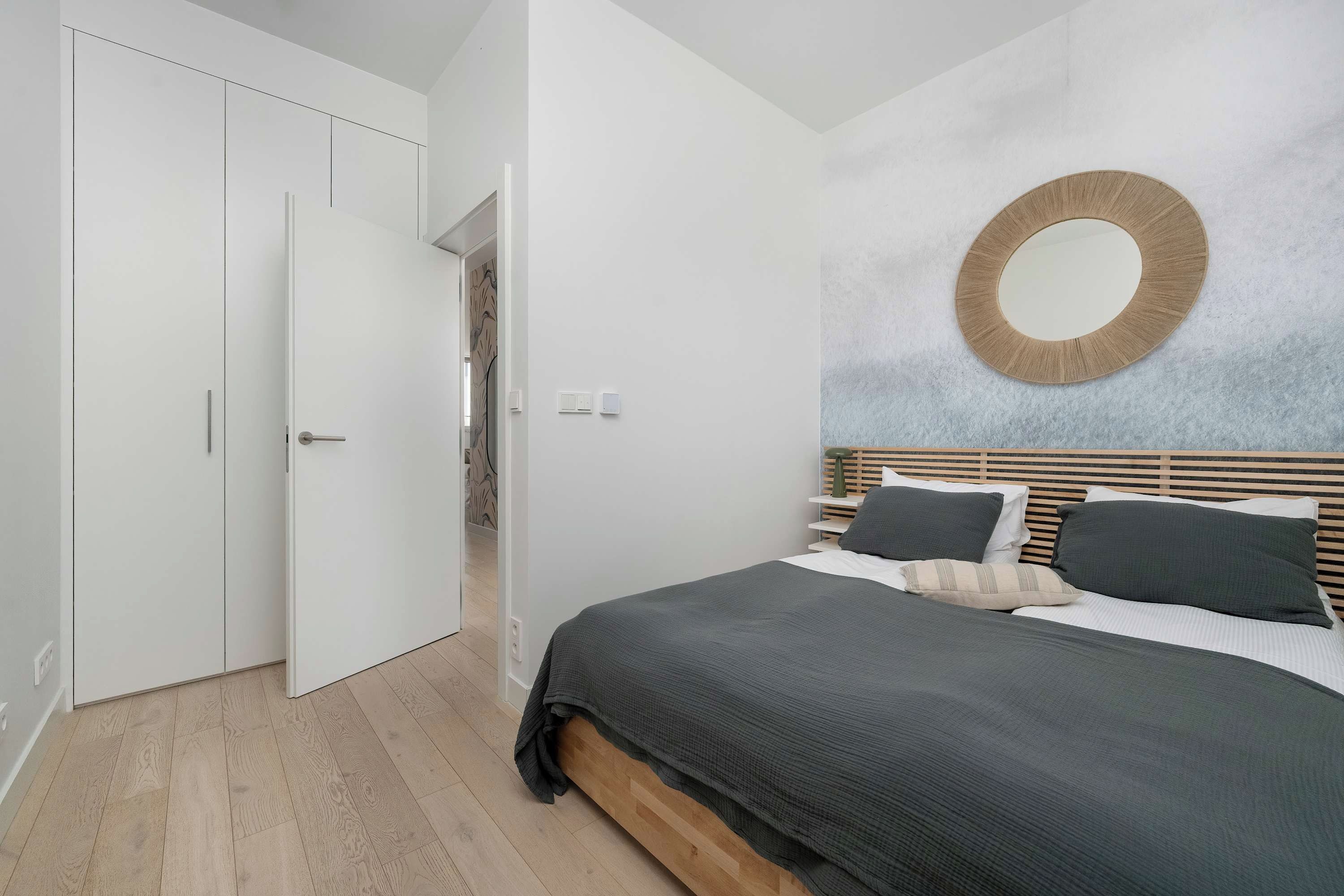 Un dormitorio con una cama, cabecera de madera, un espejo redondo y un armario blanco.