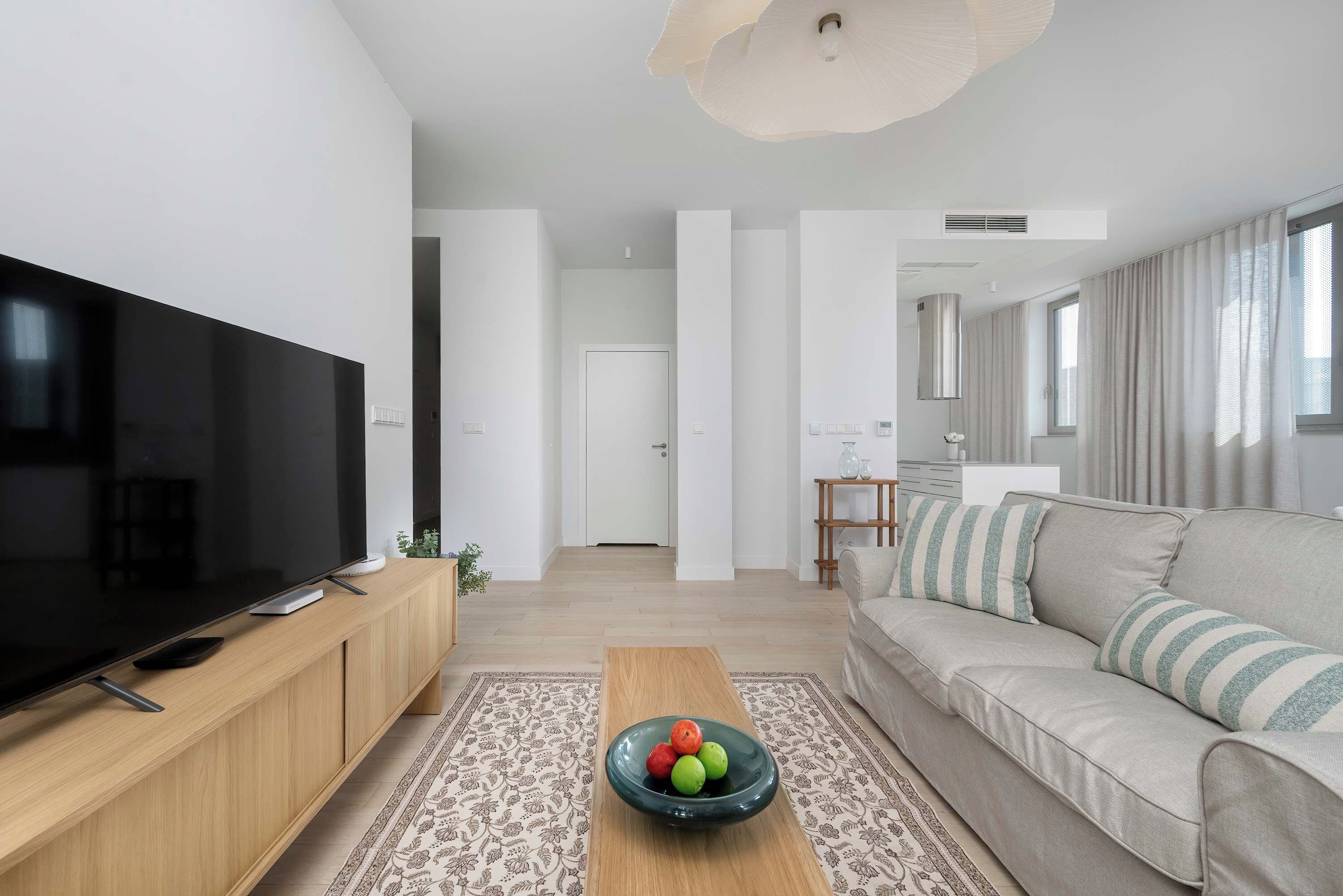Ein modernes Wohnzimmer mit einem Flachbildfernseher, einer Couch, einem Couchtisch mit Obst und einem gemusterten Teppich.