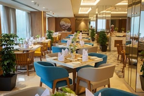 A modern dining area with tables set for service, blue upholstered chairs, and potted plants. - Arista By Ambition - Guwahati (Guwahati)