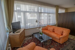 Ein Wohnzimmer mit einer Couch, einem Fernseher auf einer Holzkommode, einem Glastisch und einem großen Fenster mit durchscheinenden Vorhängen.