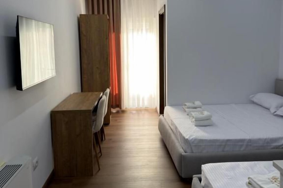 A hotel room with a wooden desk, two beds, a flat-screen TV, and a window with curtains.
