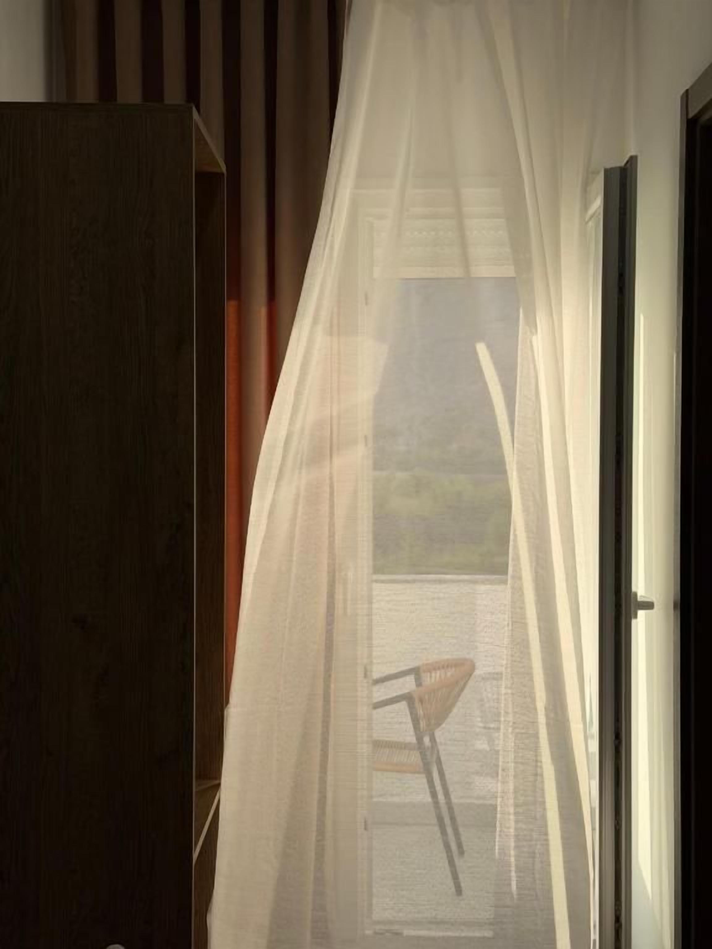 Una habitación con vistas al exterior, visible a través de una ventana con cortinas translúcidas, una puerta de madera y una silla.