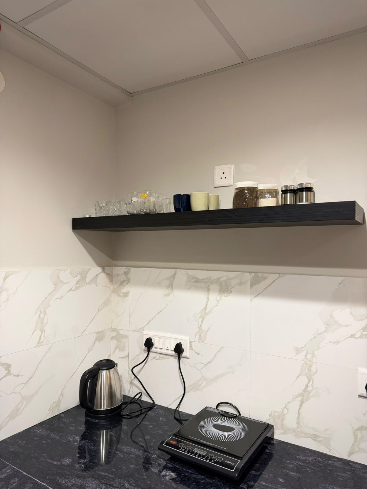 Una cocina con protector contra salpicaduras de mármol, encimera negra, hervidor, calentador portátil y un estante con diversos objetos.