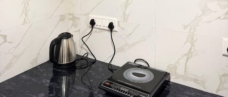 A kitchen with a marble backsplash, a black countertop, a kettle, a portable heater, and a shelf with various items.
