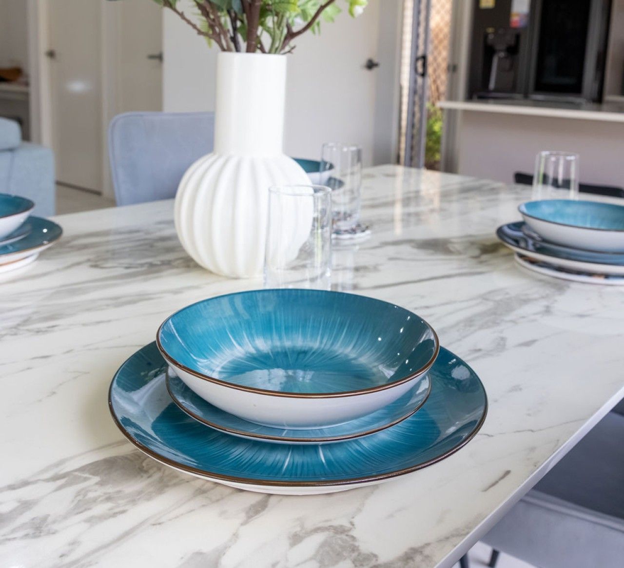 Une table à manger avec un plateau en marbre blanc, un vase blanc contenant des plantes vertes, et des assiettes bleues et blanches.