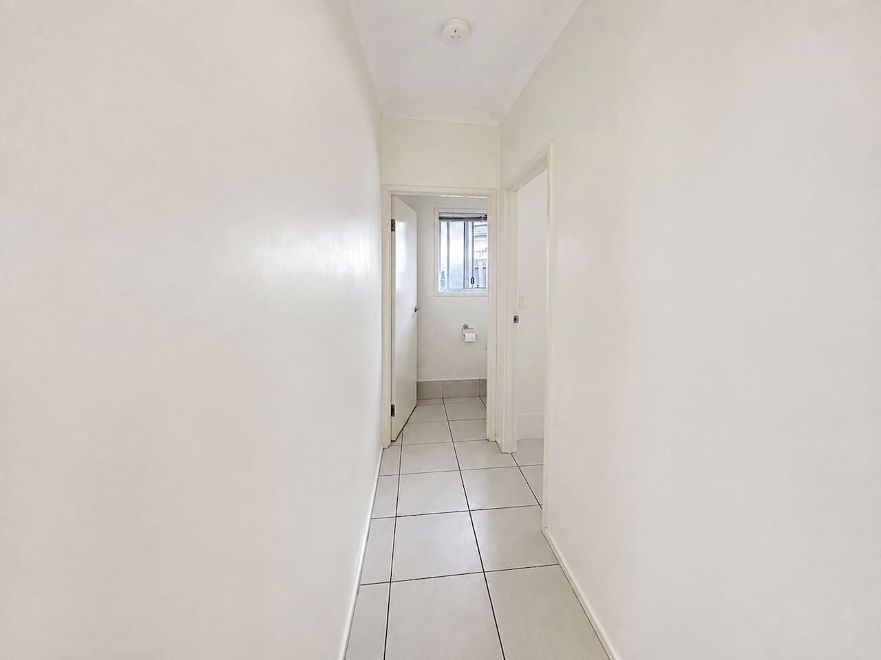 Corredor estreito com paredes brancas e chão de azulejos que leva a uma porta.