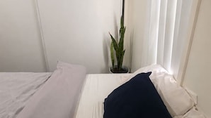 Een enkel bed met witte lakens en een donker kussen, een potplant en een raam met witte gordijnen.
