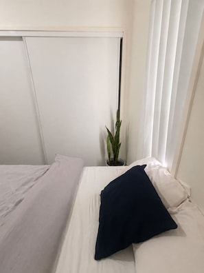 A single bed with white bedding and a dark pillow, a potted plant, and a window with white curtains. - Relaxing Gateway: Private Room 1 (North Lakes)