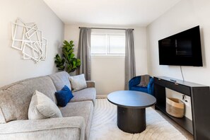 A modern living room with a grey sofa, a round coffee table, a blue chair, a large TV, and a plant. - Fremont Stylish Stay | Near K-State | Free Parking (Manhattan)