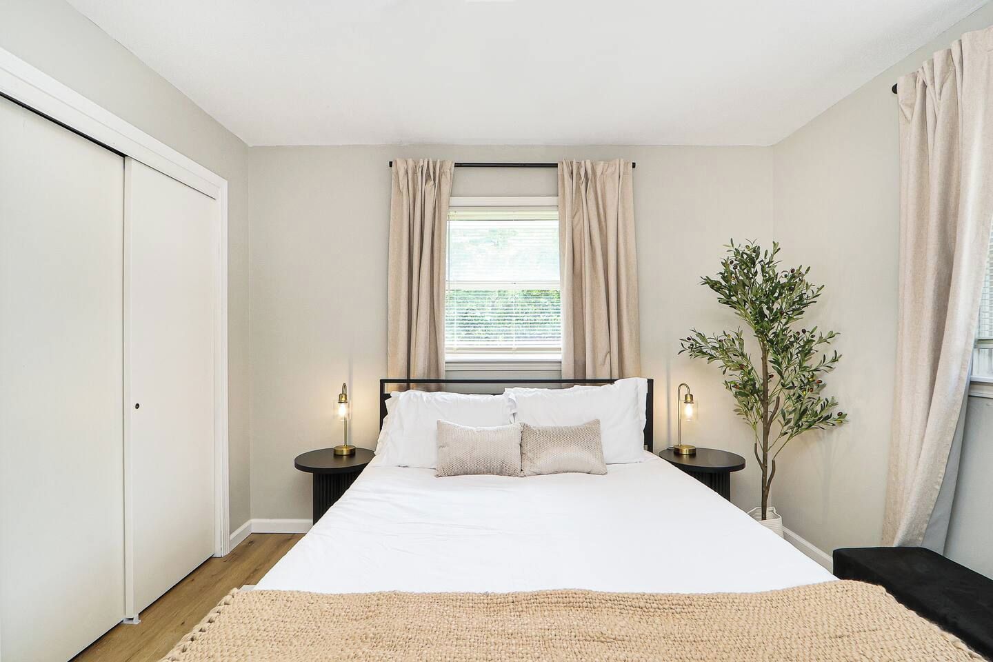 Een slaapkamer met een bed, twee nachtkastjes, een raam met gordijnen, een plant in pot en een deur.