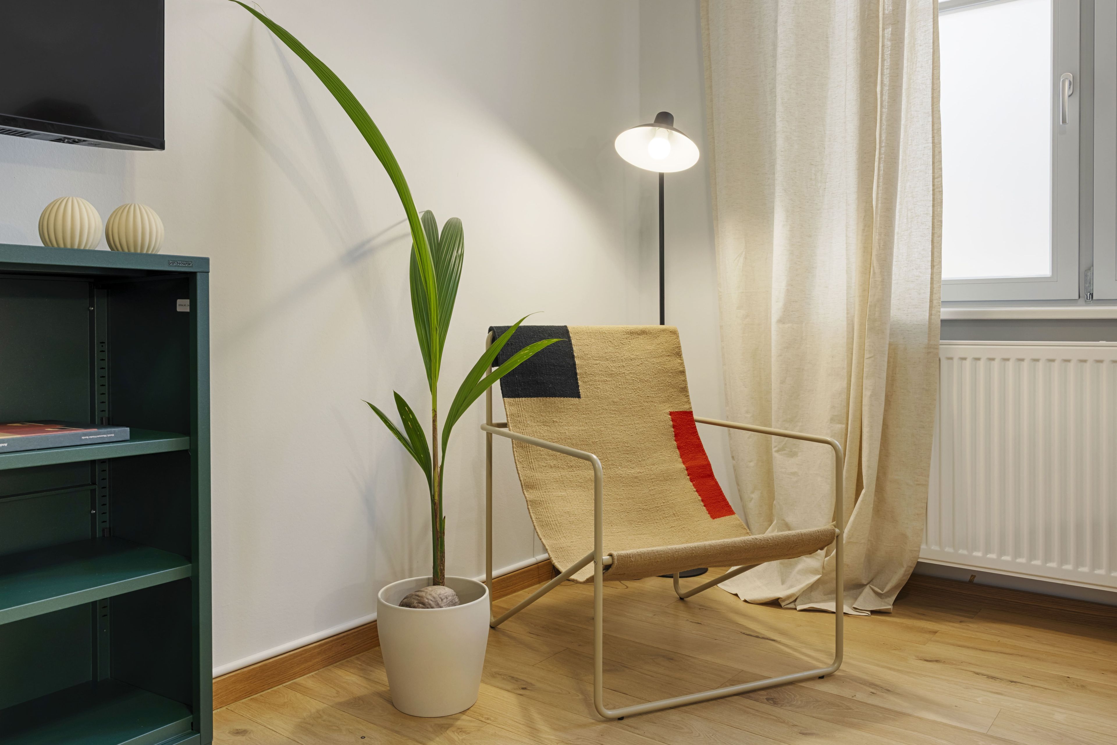 A room with a television mounted on the wall, a potted plant, a chair with a towel, a floor lamp, and a window with curtains.