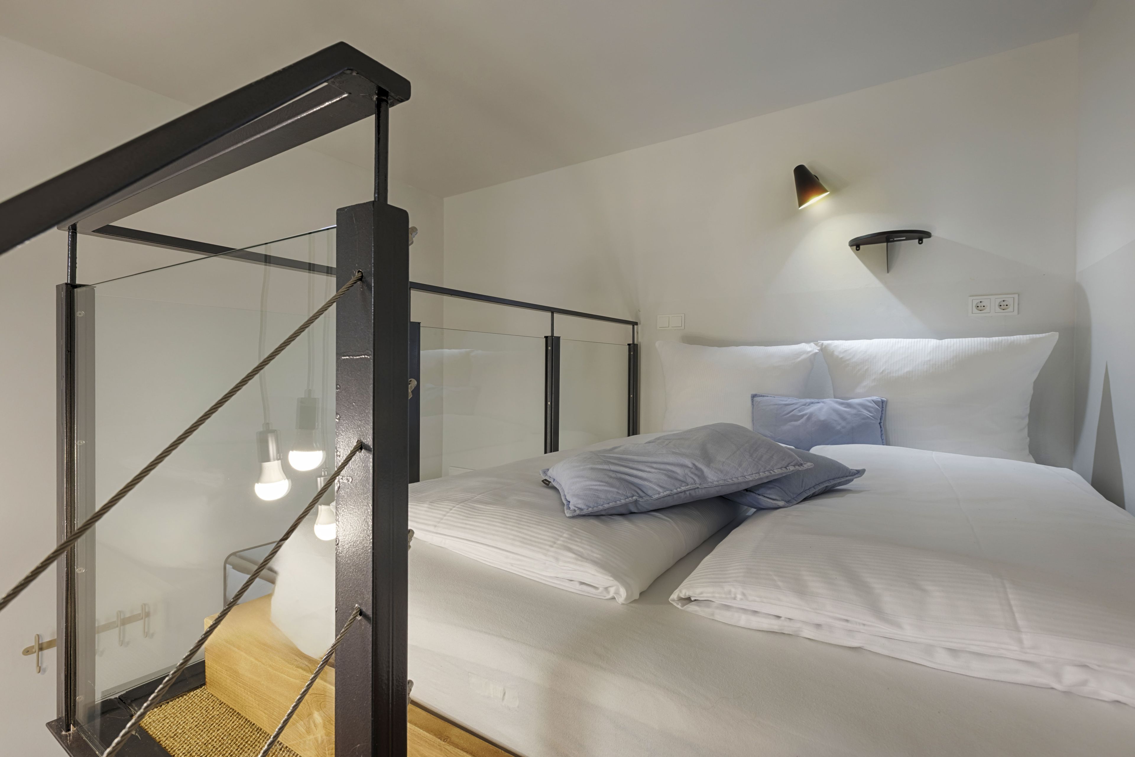 A bedroom with a bed, pillows, a headboard, a wall-mounted light, and a staircase with glass railings.