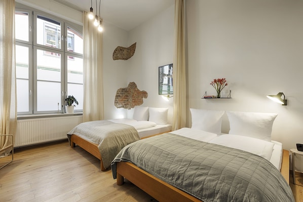 Une chambre d’hôtel avec deux lits, une grande fenêtre, un radiateur et une étagère fixée au mur, sur laquelle se trouve un vase.