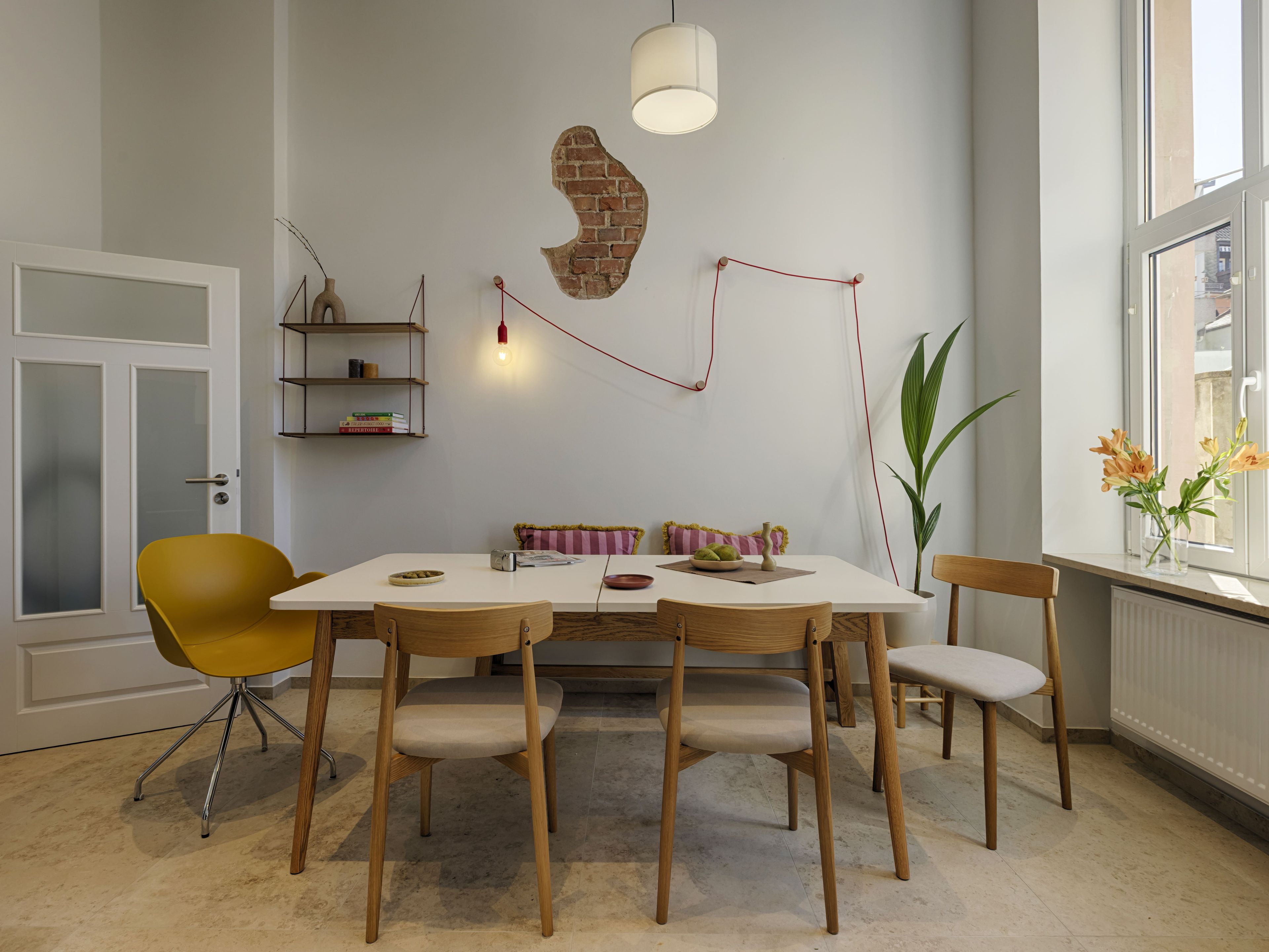 Un comedor con una mesa blanca, sillas de madera, una silla amarilla, un adorno de pared de ladrillo, una lámpara colgante y una ventana con flores.