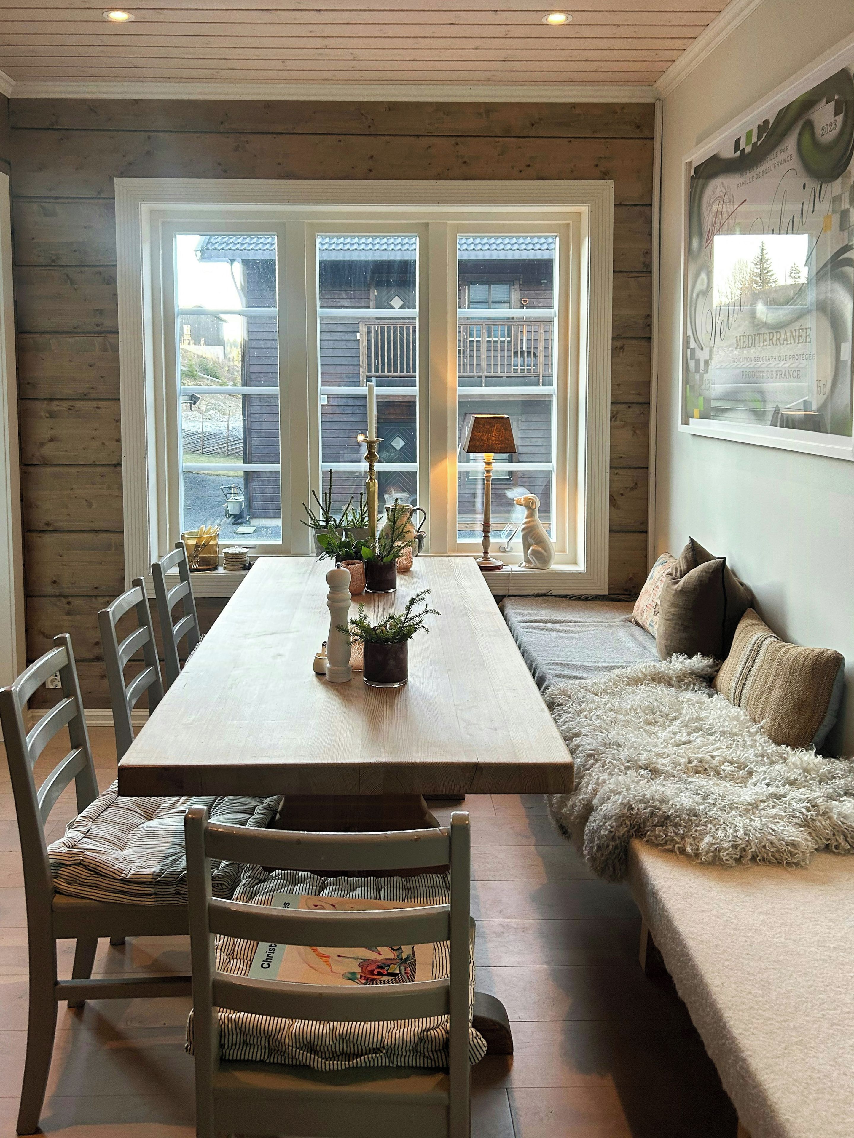 Een eetgedeelte met een houten tafel, stoelen en een bank. Er is een raam met uitzicht op een balkon en een wandlamp.