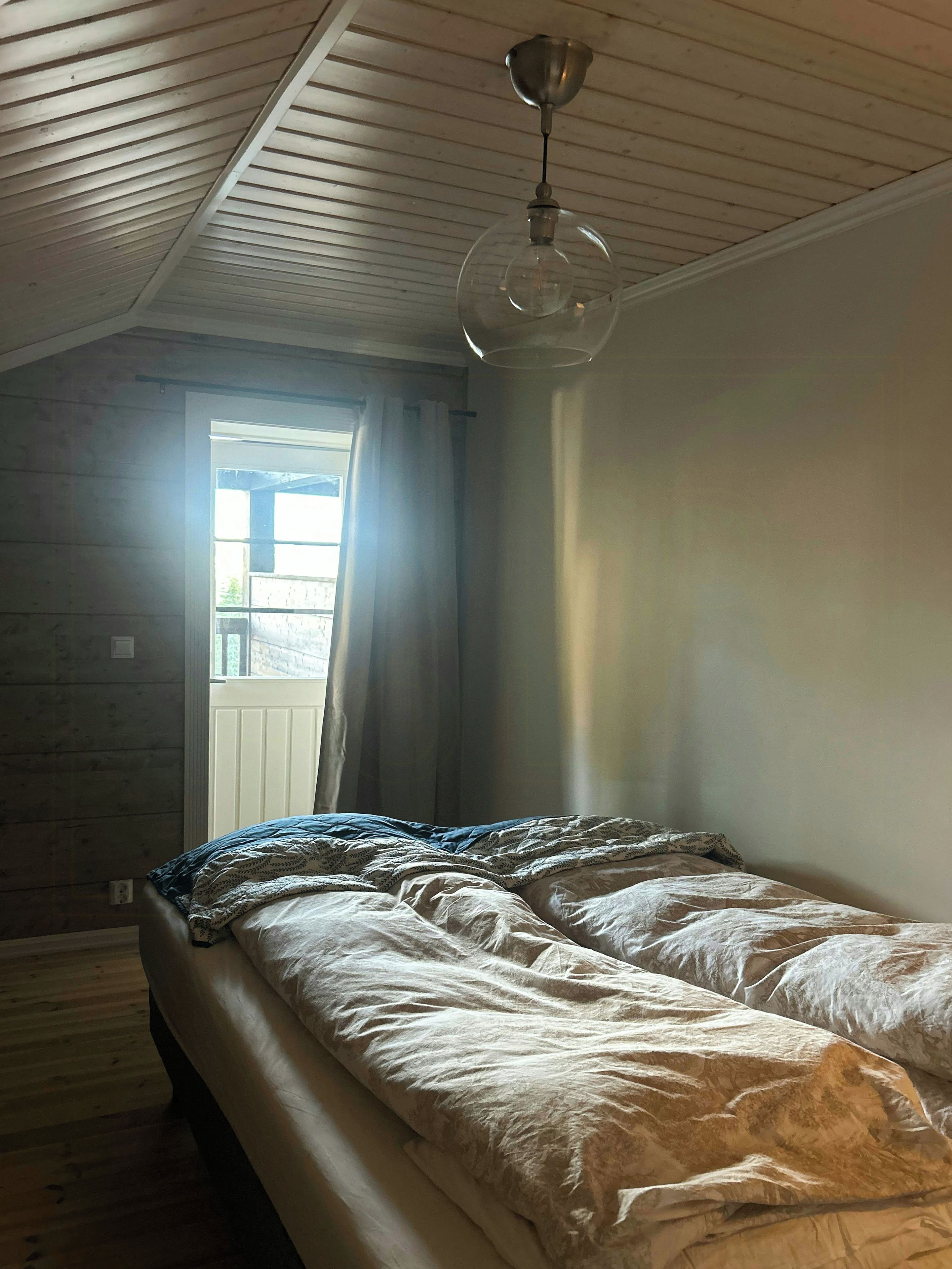 Un dormitorio con techo inclinado, una cama con edredón estampado, una ventana con cortinas y una lámpara colgante.
