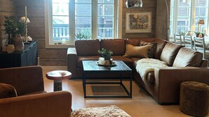 A spacious living room with a brown leather sofa, a patterned ottoman, and a coffee table. There are windows with a view of a wooden deck and a framed picture on the wall.