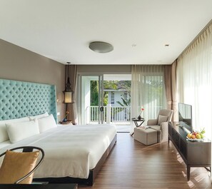 Habitación de hotel con una cama grande, zona de estar con televisor y balcón con vistas a zonas verdes.