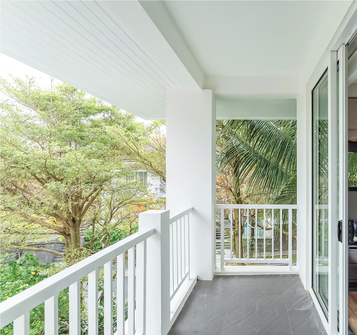 A balcony with white railings, a dark floor, and a view of trees and a building.