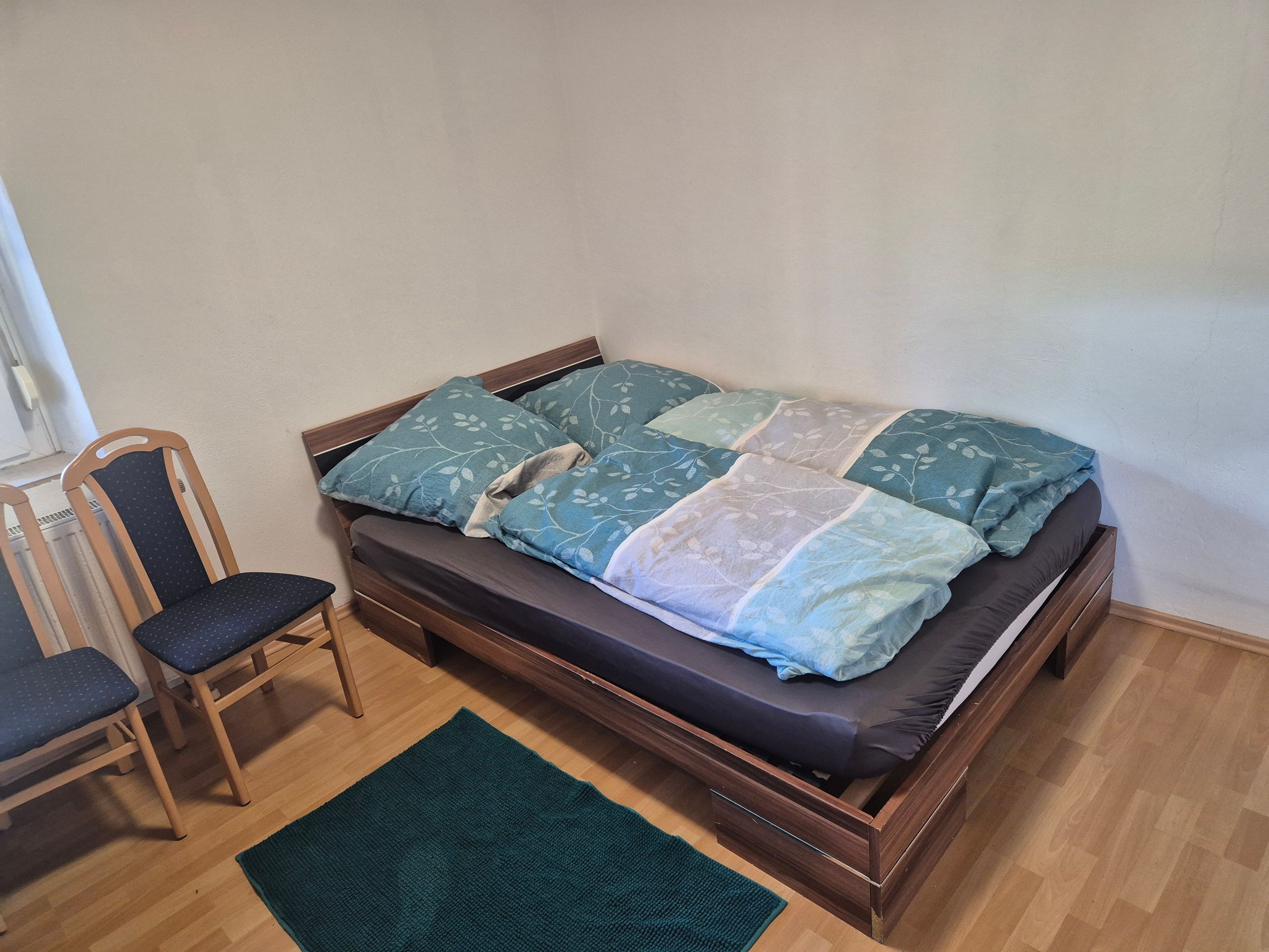 Un lit simple avec un cadre en bois, un matelas foncé et une courtepointe ornée de motifs de feuilles. Il y a un tapis vert sur le sol et une chaise en bois avec un coussin foncé.