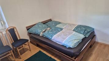 Un lit simple avec un cadre en bois, un matelas foncé et une courtepointe ornée de motifs de feuilles. Il y a un tapis vert sur le sol et une chaise en bois avec un coussin foncé.