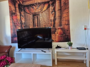 A flat-screen TV on a white stand, a white table with a plant and a router, and a wall with Egyptian-themed artwork.