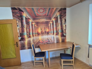 A room with a dining table and two chairs, a mural on the wall, and a wooden door.
