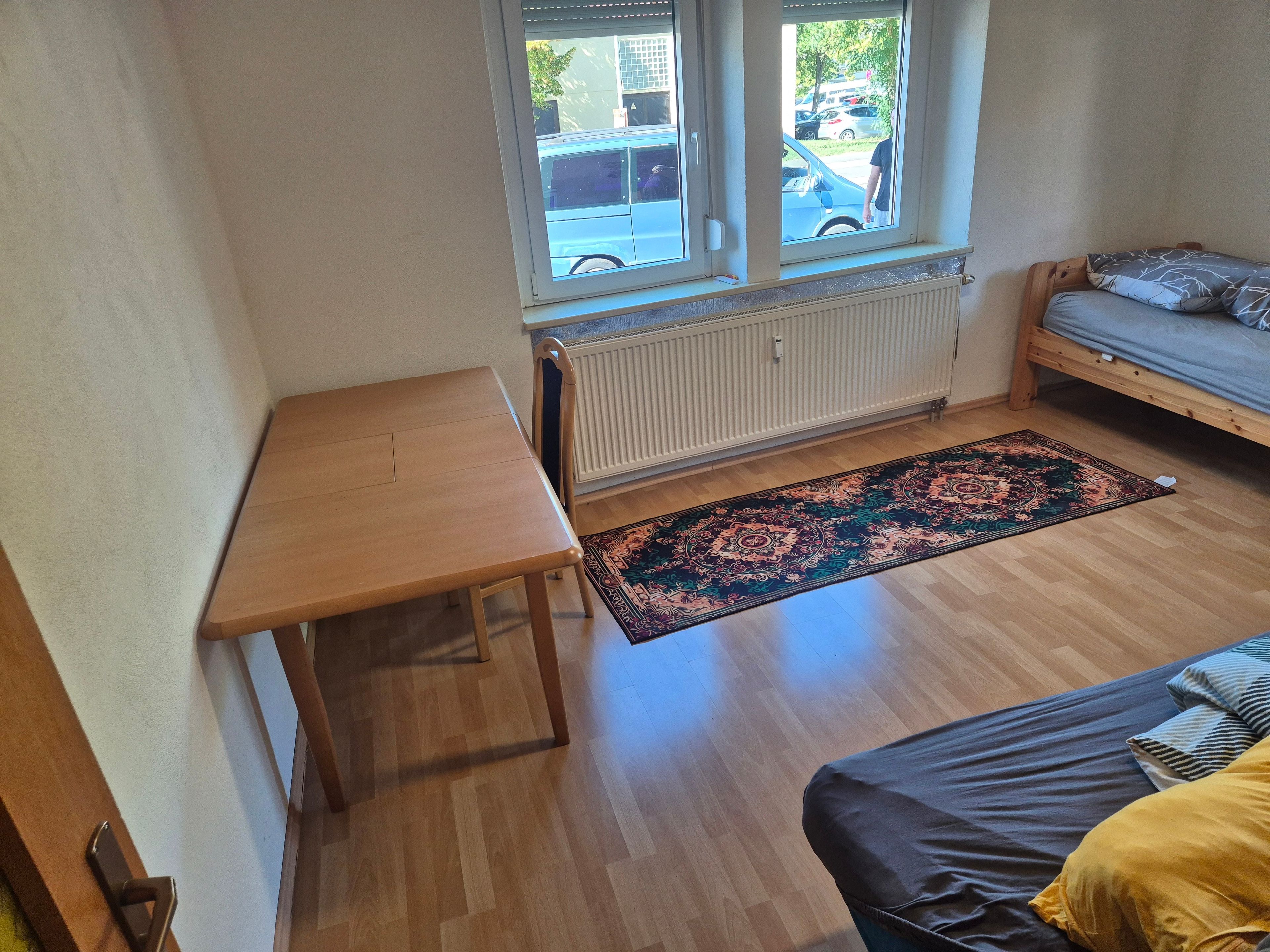 Een kamer met een houten tafel, een bed met een grijze matras en gestreepte kussens, een raam met uitzicht op geparkeerde auto's, en een vloerkleed met een patroon.