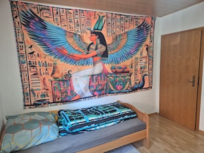 Una stanza con un letto, una porta in legno e un arazzo raffigurante geroglifici egizi e una figura alata.