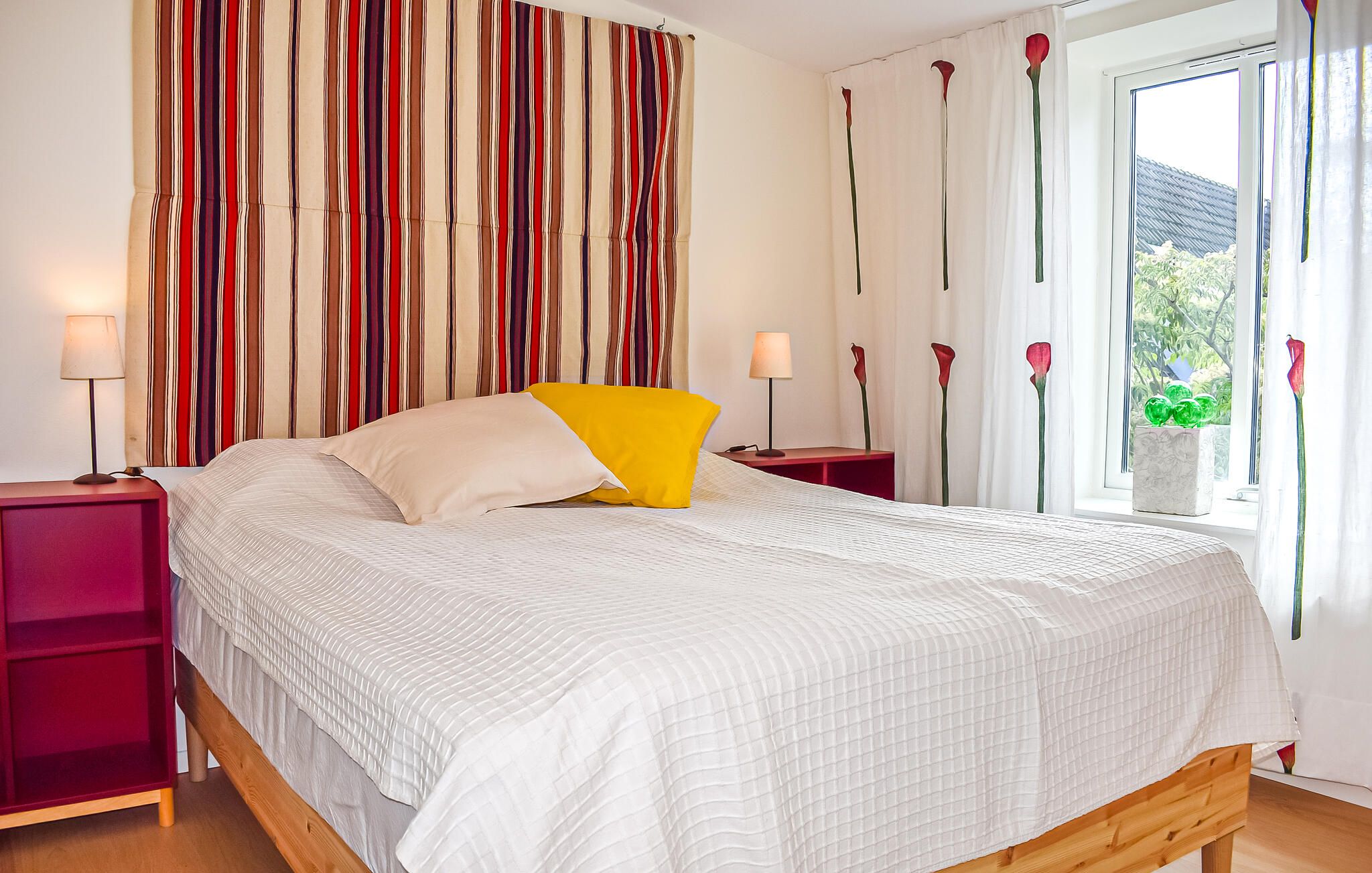 A bed with a white quilted bedspread and a yellow pillow, flanked by two nightstands with lamps, in a room with a striped curtain backdrop and a window with a view of greenery.