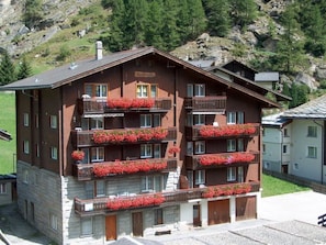 A multi-story building with balconies adorned with red flowers, set against a mountainous backdrop. - Bergfreude Kuonen Appartement/Fewo 4-Bettwohnung by Interhome (Saas-Grund)