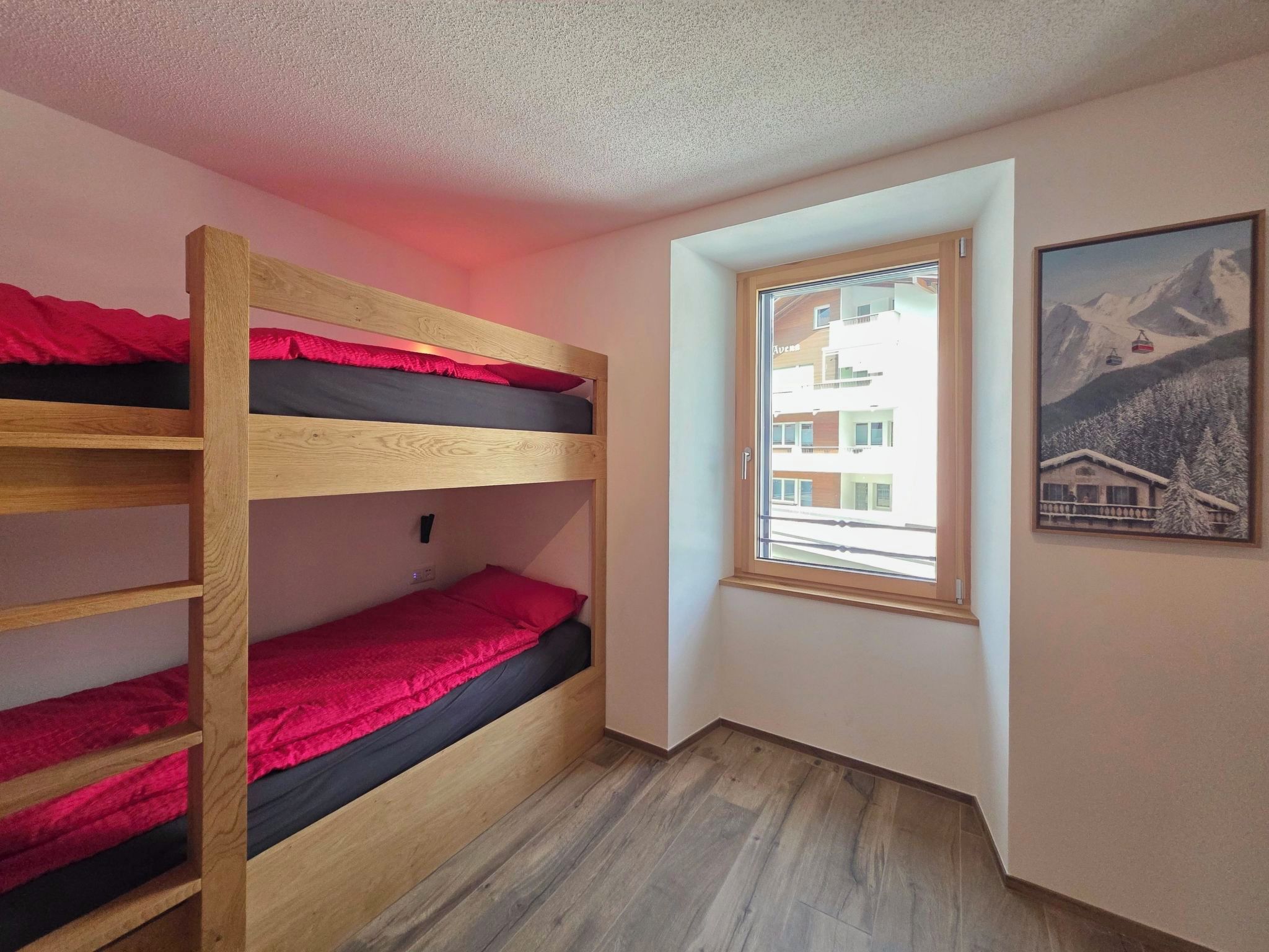 Une chambre avec un lit superposé, une fenêtre donnant sur des immeubles et un tableau encadré accroché au mur.