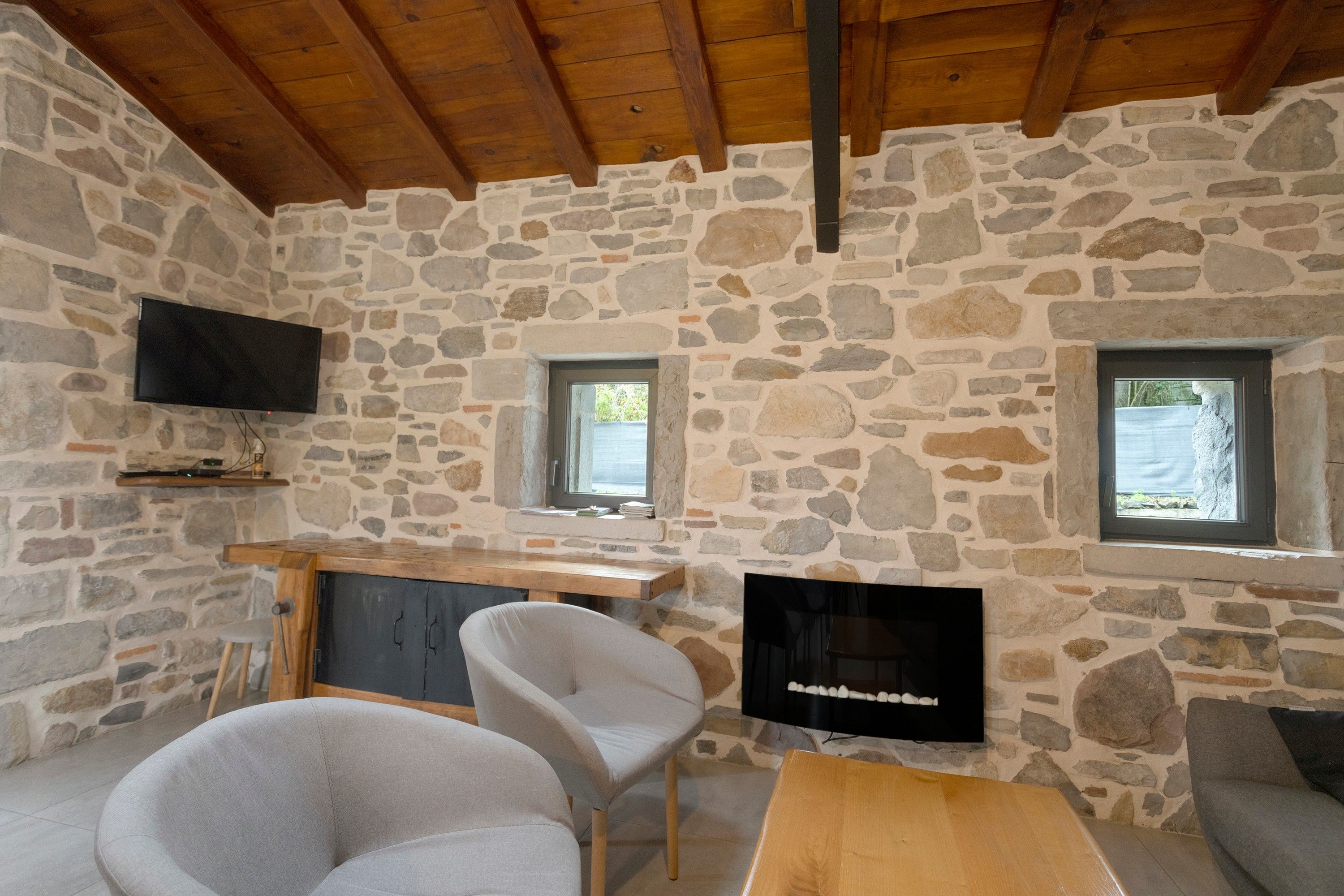 A stone-walled room with a wooden ceiling, a flat-screen TV mounted on the wall, a wooden table, two chairs, and a sofa.