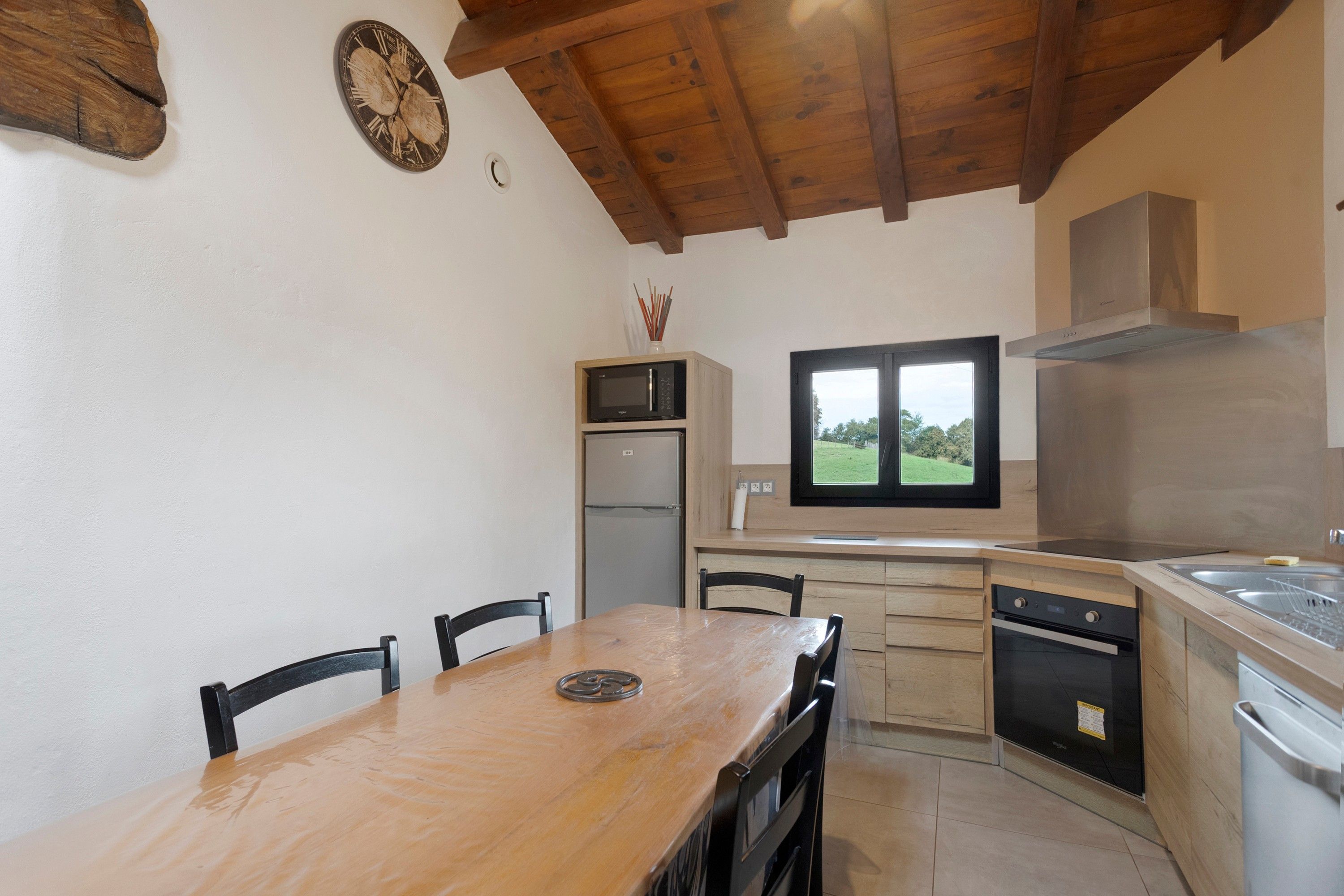 A compact kitchen with a wooden table, chairs, a built-in oven, a microwave, and a window with a view of greenery.