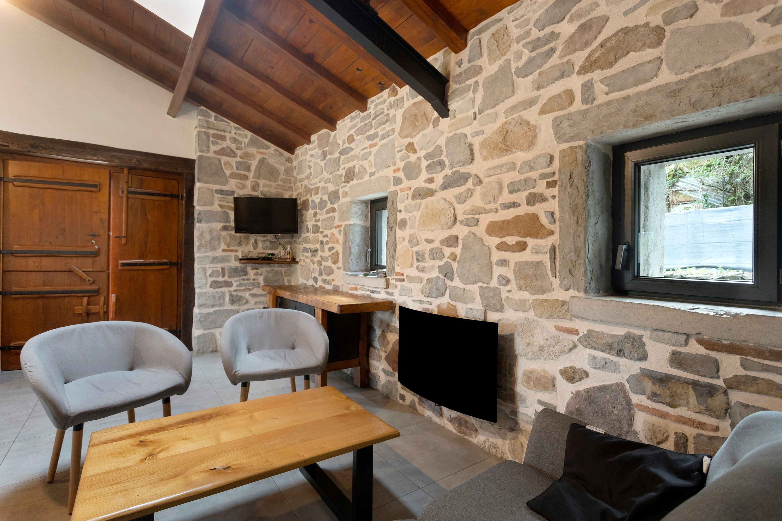 A stone-walled room with a wooden bench, two chairs, a TV mounted on the wall, and a window with a view.
