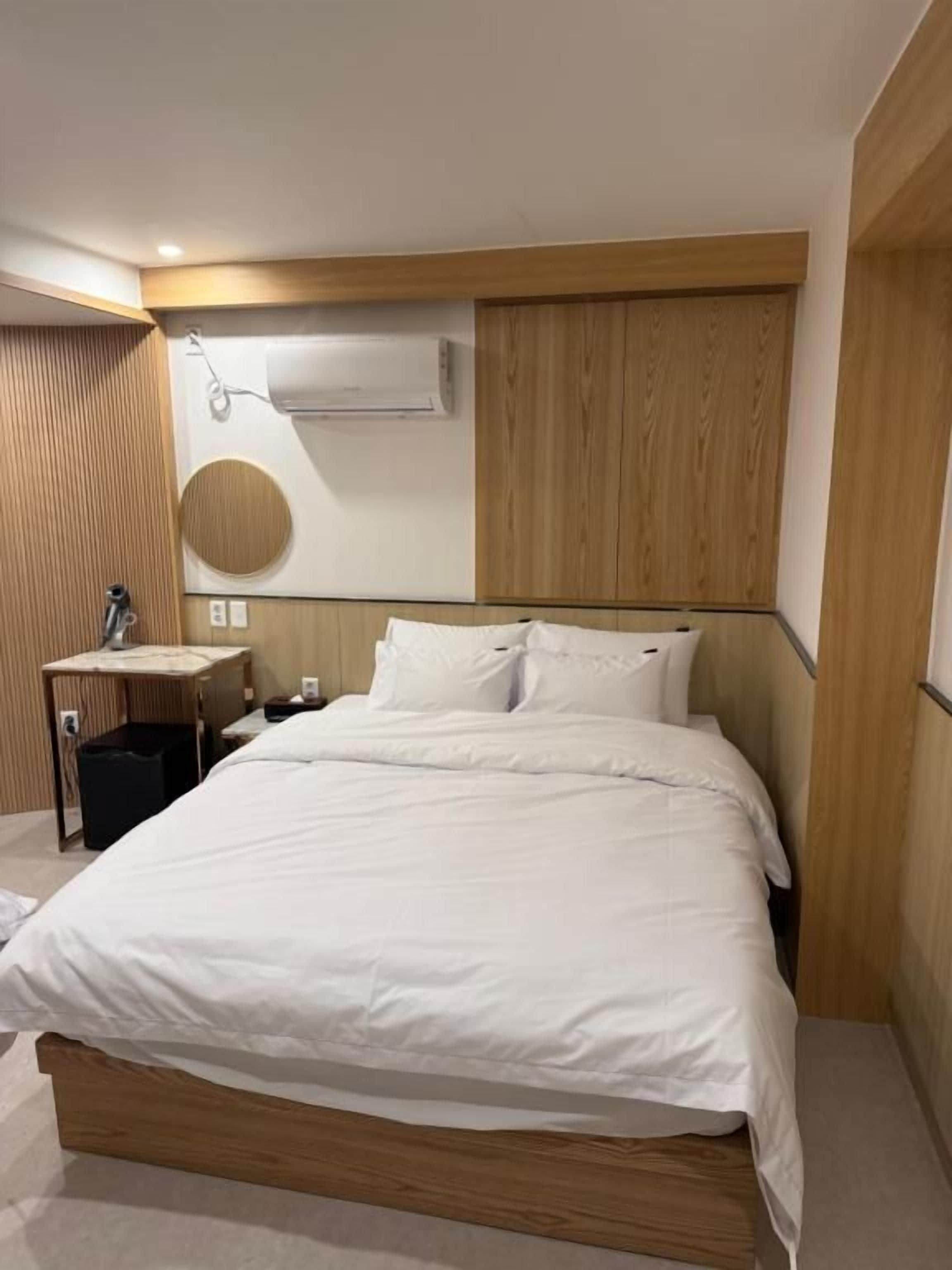 A hotel room with a large bed, a bedside table, a wall-mounted air conditioner, and a circular mirror.